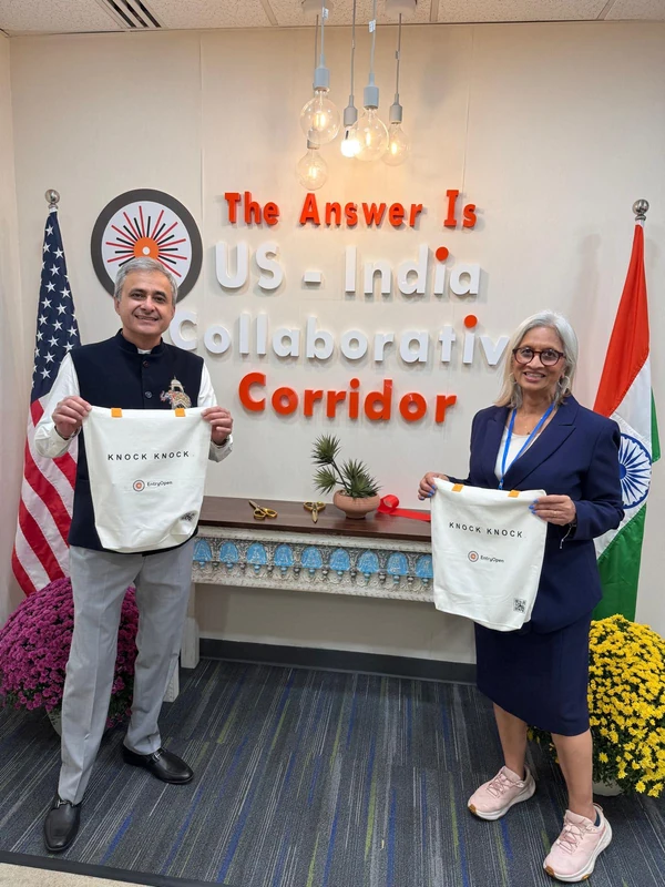 Two guests hold EntryOpen welcome bags in front of the U.S.–India Collaborative Corridor wall and the Indian and American flags.